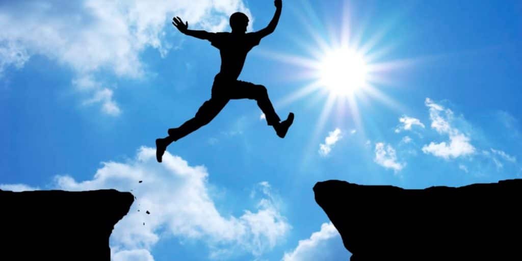 Silhouette of a man jumping over a gap between two cliffs while the sun shines in a blue, cloudy sky.