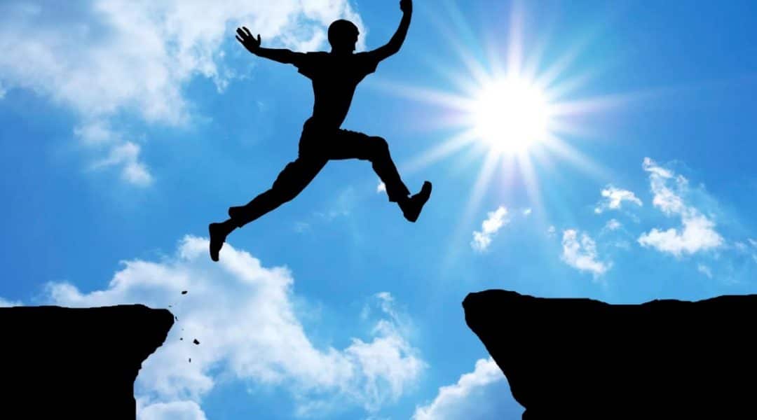 Silhouette of a man jumping over a gap between two cliffs while the sun shines in a blue, cloudy sky.