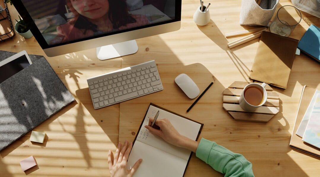 A woman at a desk writes in a notebook while the image of a woman appears on a computer monitor.