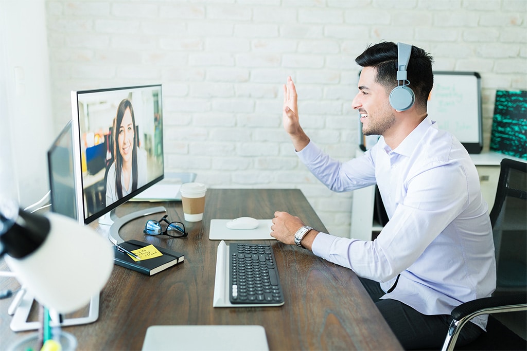 Man sitting at desk waving to woman on computer screen during video meeting.
