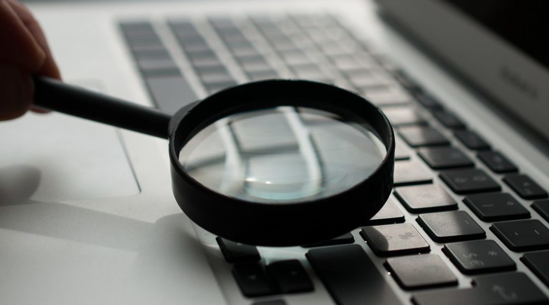A hand holds a magnifying glass over a laptop keyboard.