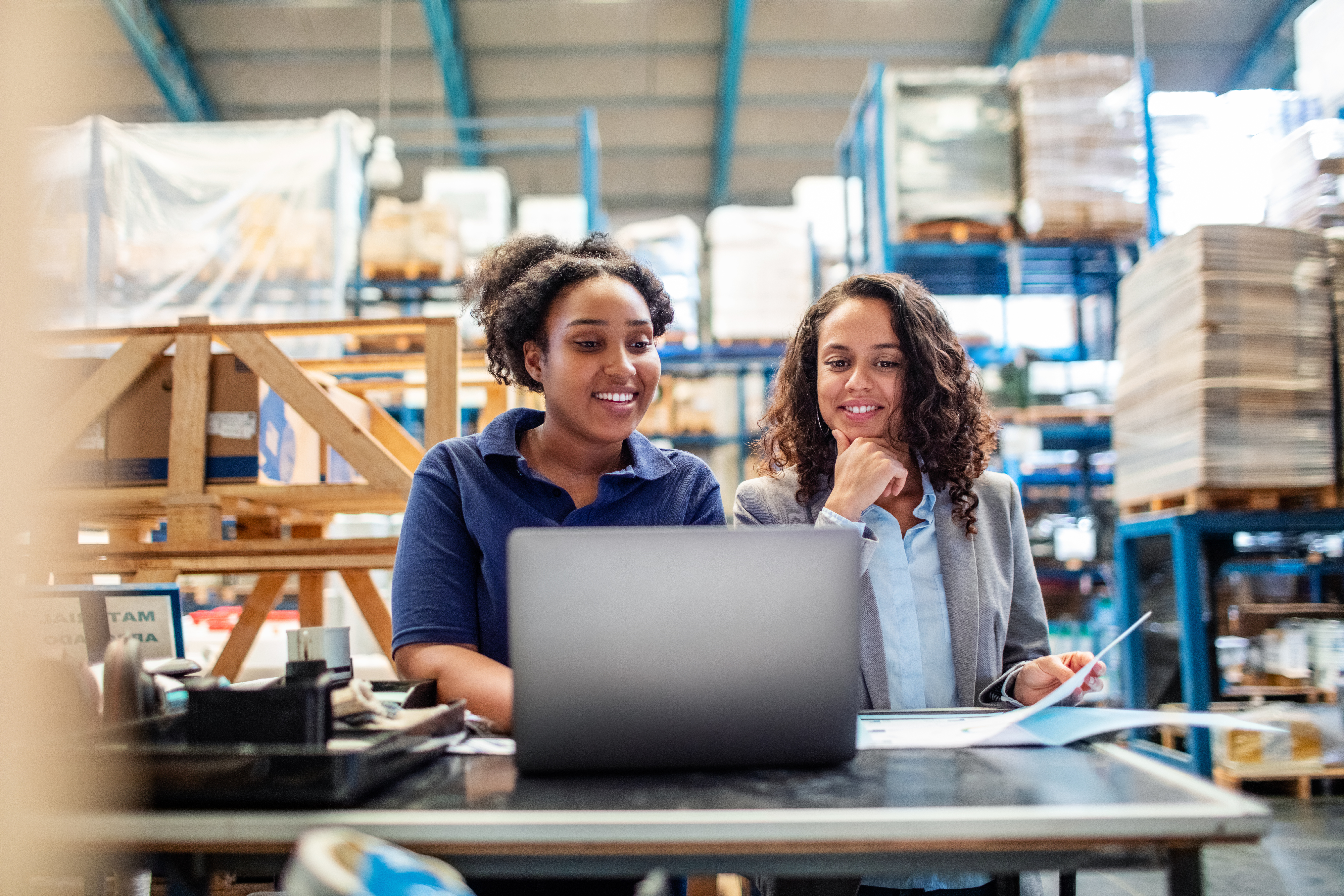Two women look at a laptop screen in a warehouse.