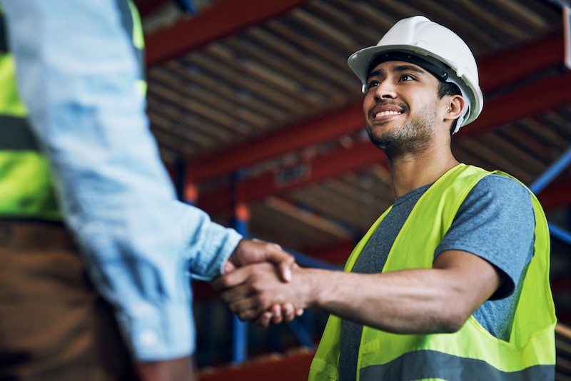 Two warehouse workers wearing hard hats and high-visibility vests shake hands.