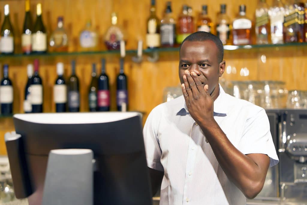 Bar tender is standing at a cash register with his hand on his mouth